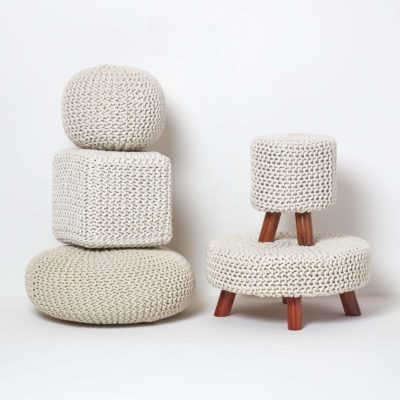 Two stacks of knitted pouffes. On the left, a flat oval pouffe, tall cube pouffe and small round pouffe. On the right, a short oval pouffe with wood legs, and a taller cylinder pouffe with legs.